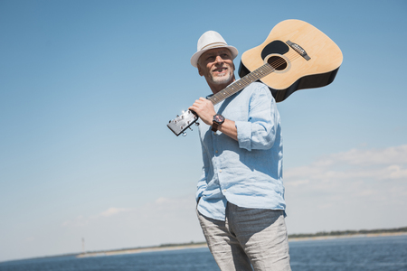 senior man in hat holding acoustic guitar on shoulder and looking away outdoorsの写真素材
