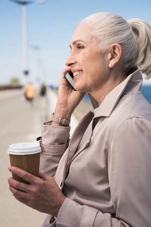 senior woman holding disposable coffee cup and talking on smartphoneの写真素材