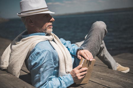 senior man lying with book and looking away on quayの写真素材