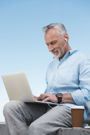 senior man typing on laptop and listening music in earpods outdoorsの写真素材