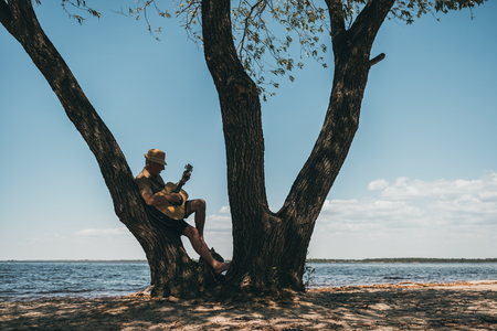 man in hat leaning at big tree and playing guitar at riversideの写真素材