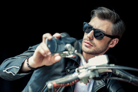 stylish young man in sunglasses and leather jacket sitting on motorcycle and looking awayの写真素材