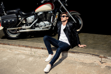 stylish young man in leather jacket and sunglasses sitting on concrete curb near motorbikeの写真素材