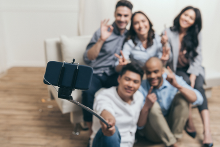 smiling multiethnic friends taking selfie with monopod and smartphone at homeの写真素材
