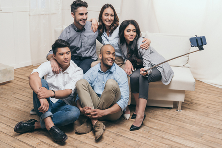 smiling multiethnic friends taking selfie with monopod and smartphone at homeの写真素材