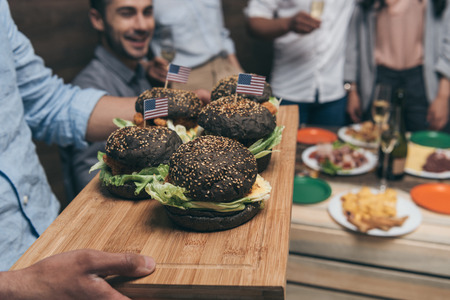 person holding wooden tray with delicious hamburgers and friends partying behindの写真素材