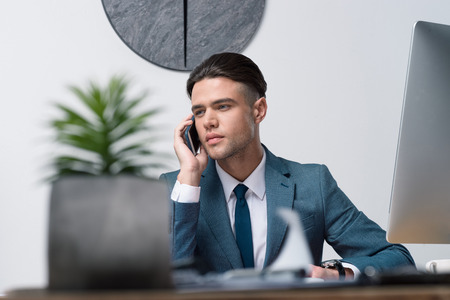 young businessman talking on smartphone and looking away in officeの写真素材