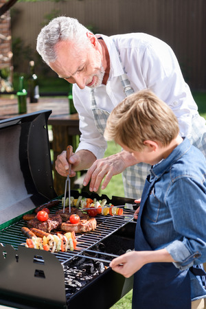 haired grandfather with his grandson preparing meat and vegetables on grill outdoorsの写真素材
