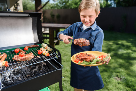 kid boy in apron preparing tasty stakes on barbecue grill outdoorsの写真素材