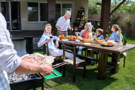 man holding tray with meat while family sitting at table outdoorsの写真素材