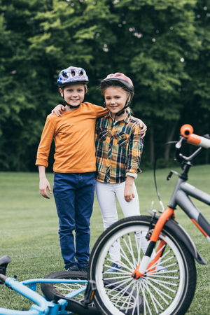 kids in helmets embracing and looking at camera while standing near bicycles at parkの写真素材