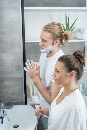 man shaving his beard while woman brushing teeth in bathroomの写真素材