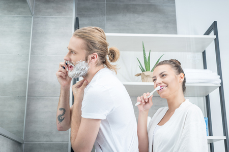 young man shaving beard with razor and smiling woman brushing teeth in bathroomの写真素材