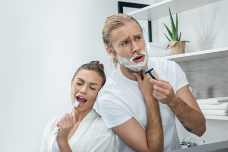 young man shaving beard with razor while happy woman singing in toothbrushの写真素材