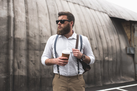 bearded man with sunglasses and leather backpack holding disposable coffee cupの写真素材