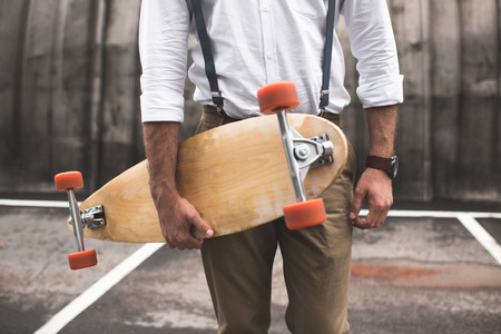 cropped view of fashionable man in white shirt and suspenders holding longboardの写真素材
