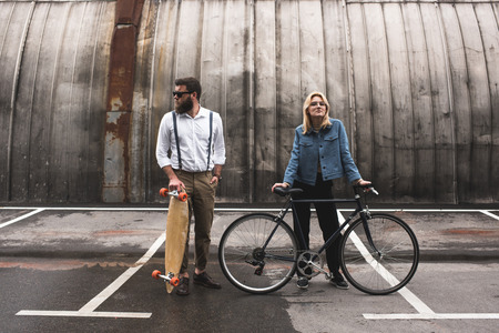 young couple standing with bicycle and longboard on parking place