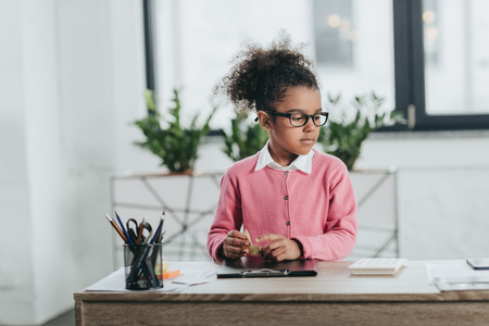 little girl in eyeglasses sitting at table with office suppliesの写真素材