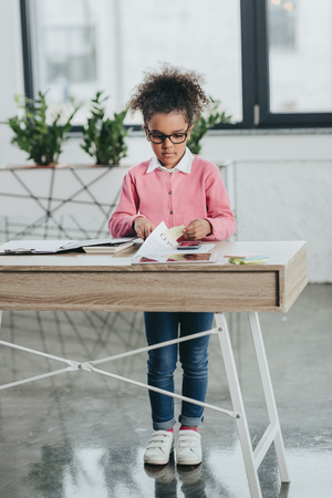 little girl in eyeglasses cutting paper sheet with scissors while standing at office tableの写真素材