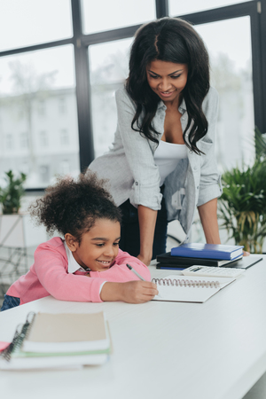 mother looking at little daughter writing in notebook at workplaceの写真素材