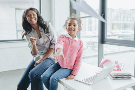 daughter with her mother throwing paper airplane while sitting at tableの写真素材