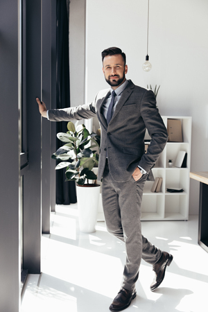 bearded businessman in suit posing with hand in pocket at modern officeの写真素材