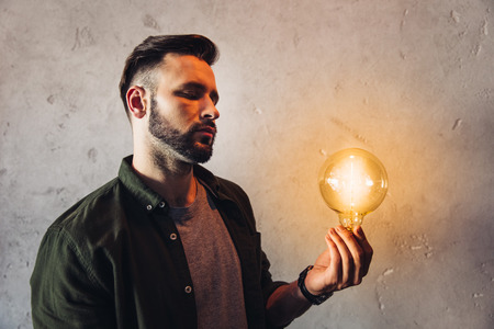 bearded man holding illuminated light bulb while standing near grey wallの写真素材