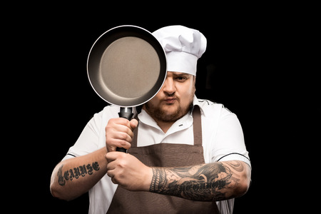 young chef in hat and apron holding frying pan isolated on blackの写真素材