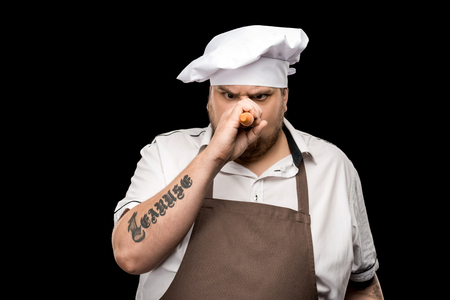chef in hat and apron holding raw carrot near nose and looking at cameraの写真素材