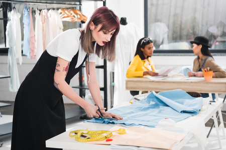 dressmaker cutting blue fabric with scissors while her colleagues talking on backgroundの写真素材