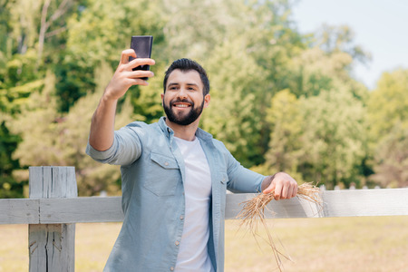 bearded man standing at wooden fence and taking selfie on smartphoneの写真素材