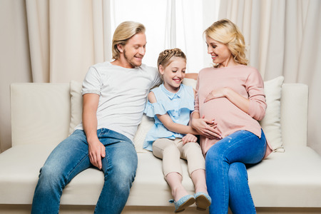 pregnant woman with smiling husband and daughter sitting on sofa at homeの写真素材