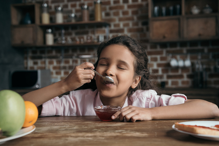 little girl with eyes closed eating sweet jam from teaspoonの写真素材