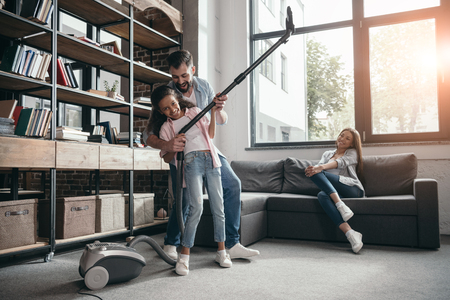 father with daughter holding vacuum cleaner while mother sitting at sofaの写真素材
