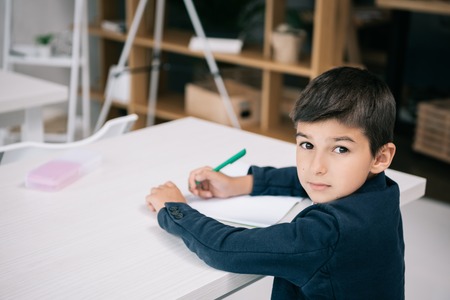 serious schoolboy writing in exercise book while sitting at desk and looking at camera