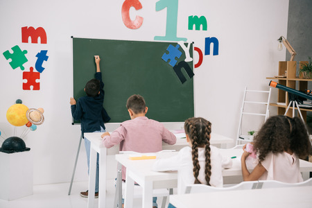 schoolboy writing on chalkboard while classmates studying at desksの写真素材