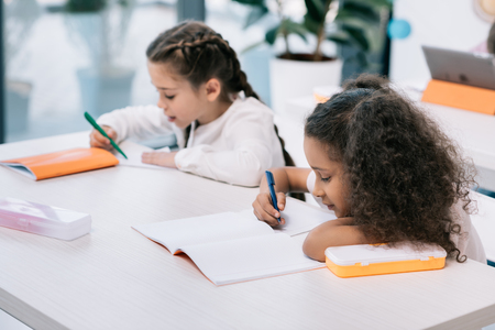 Adorable multiethnic schoolgirls writing and studying at desk in classの写真素材