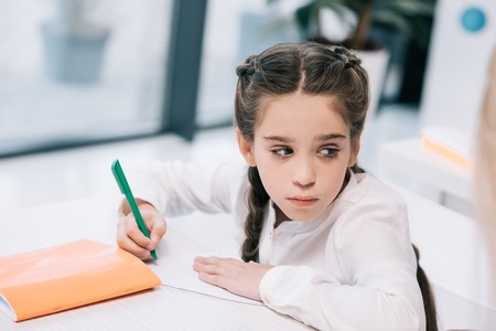 pensive schoolgirl holding pen and looking away in classroomの写真素材