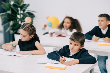 little schoolchildren sitting at desks and writing in exercise booksの写真素材