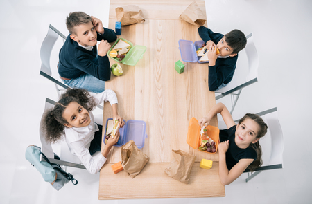 multiethnic schoolkids eating lunch while sitting at table and smiling at cameraの写真素材