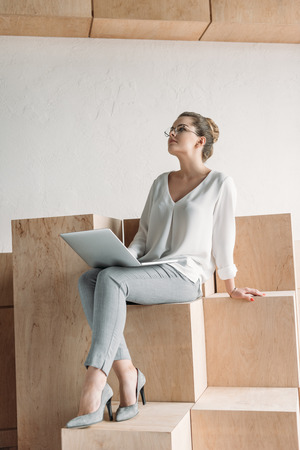 pensive fashionable businesswoman working with laptop while sitting on wooden cubesの写真素材