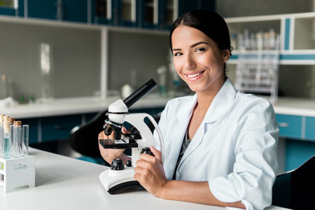 young chemist in white coat working with microscope and smiling at camera in labの写真素材