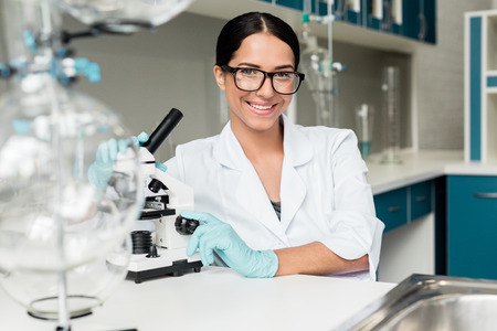 young scientist in eyeglasses working with microscope and smiling at camera in chemical labの写真素材