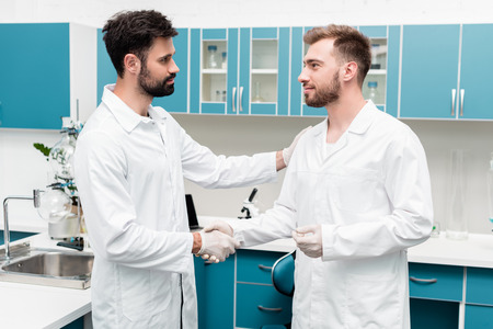 young chemists in white coats shaking hands in scientific laboratoryの写真素材
