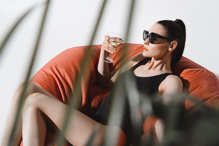 young woman in sunglasses and bodysuit drinking cocktail while resting in bean bag chair