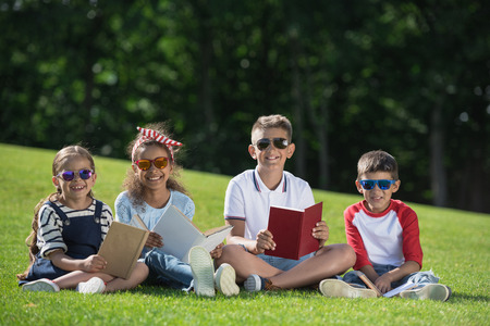 smiling multiethnic kids in sunglasses holding books and smiling at camera in parkの写真素材