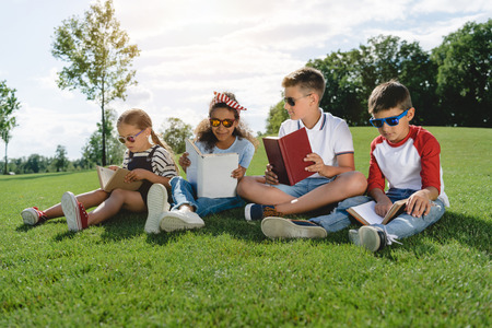 multiethnic children in sunglasses reading books on green meadowの写真素材