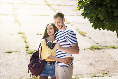 young multiethnic couple of tourists holding paper cup and camera while hugging outdoorsの写真素材