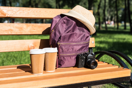 backpack with straw hat, camera and paper cups on wooden bench in parkの写真素材