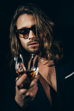 bearded long haired man in sunglasses and stylish suit drinking whiskey and looking at cameraの写真素材